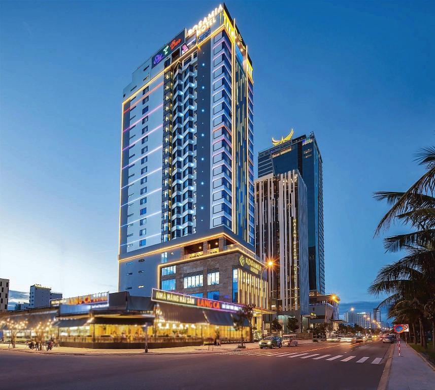 Rosamia Da Nang Hotel tower at dusk, glass facade lit, with the street and trees in front
