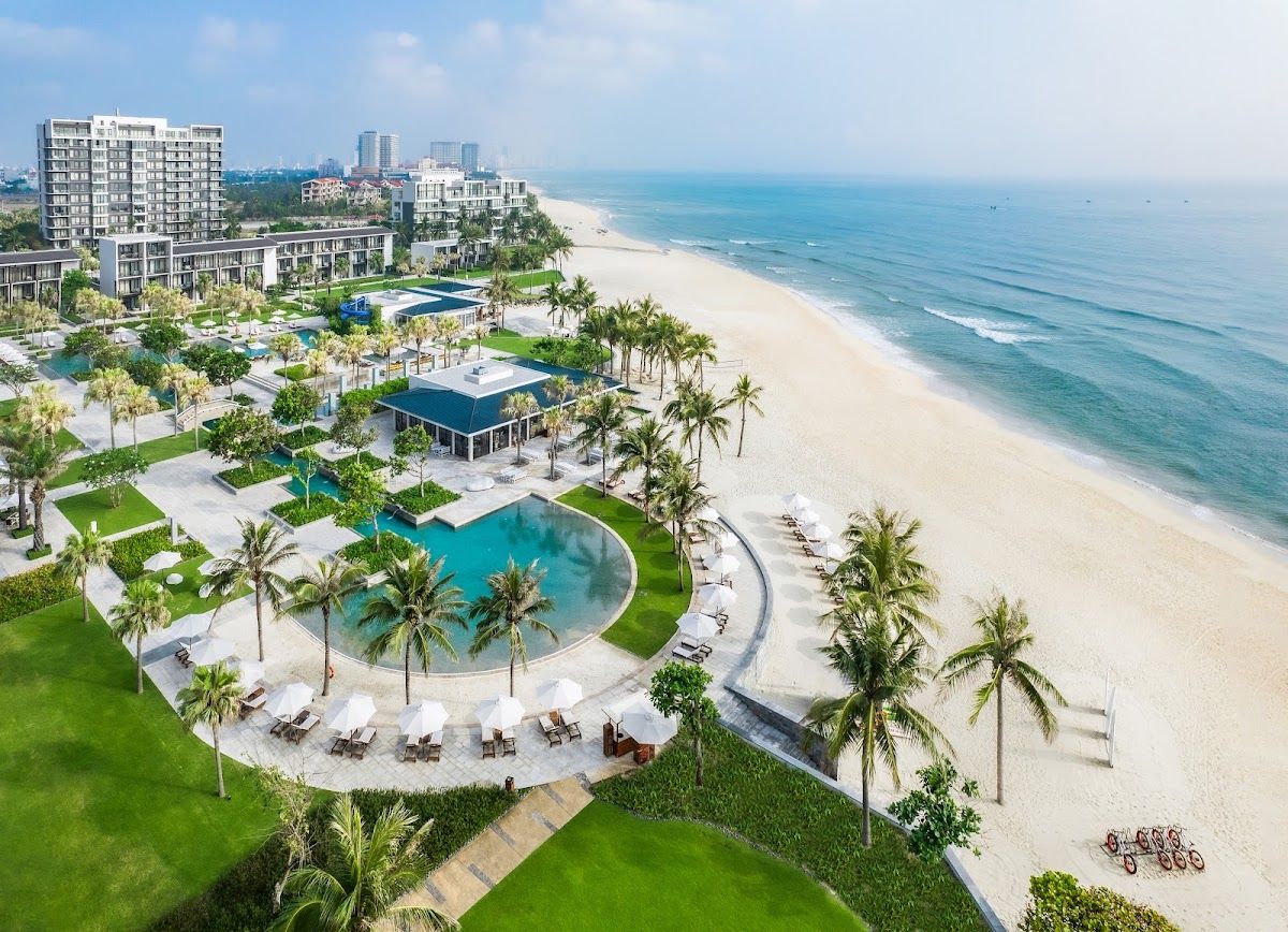 Aerial view of a luxury beachfront resort on Non Nuoc beach in Da Nang showing a curved pool, palm trees, sun loungers, and the white sand shoreline
