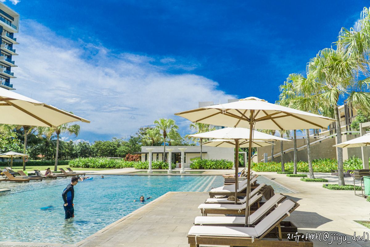 Outdoor pool at Hyatt Regency Danang with sun loungers, umbrellas, palm trees, and guests swimming under a blue sky