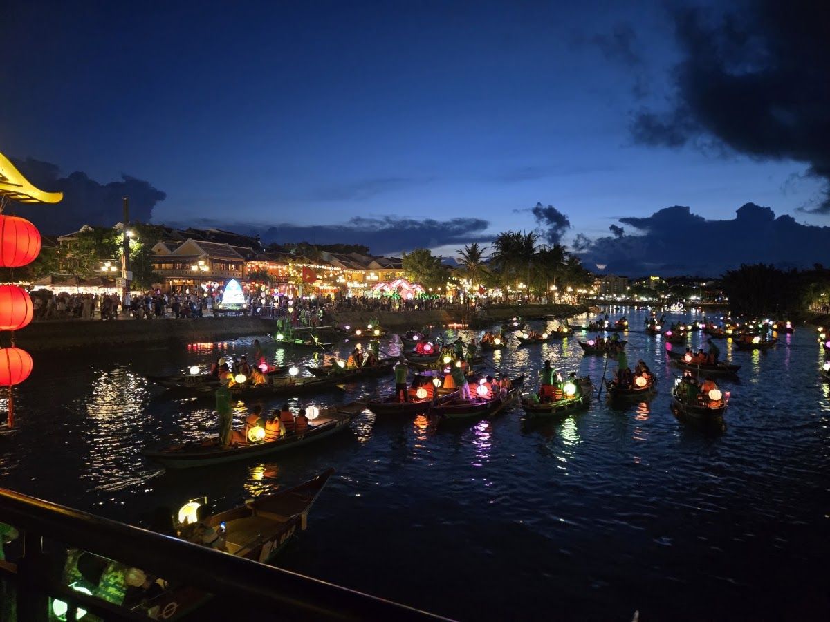 View from the Thu Bon River toward Hoi An's Old Town waterfront, with wooden fishing boats moored in the foreground and the lantern-lit facades of the Ancient Town behind them